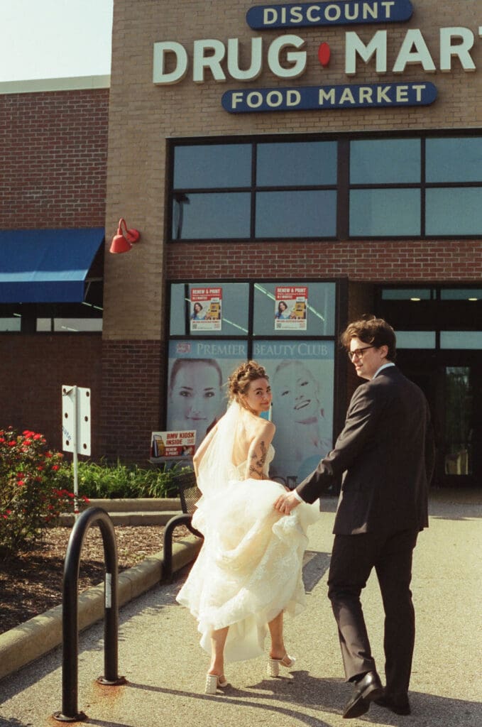 An editorial film photo of a bride and groom walking into a drug store together. the groom is holding the brides dress as they both look back at the camera