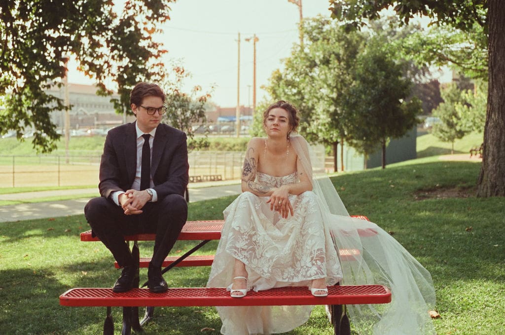 An editorial film photo of a bride and groom sitting on a park bench with a more serious look
