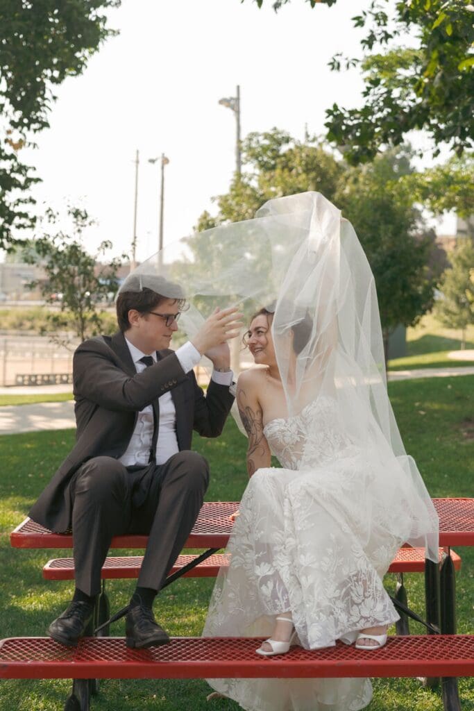 A bride and groom sitting on a playground table outside, bride is playing with her veil and letting it flow in the wind
